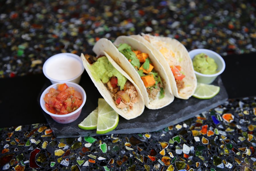 Three tacos in flour tortillas sitting between bowls of condiments on a multicolored countertop.