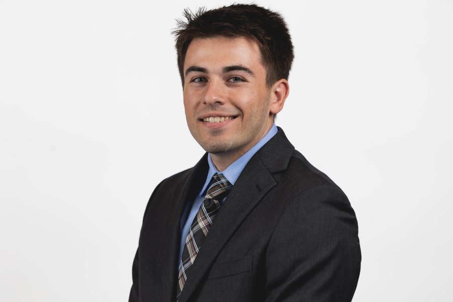 Headshot of Grant Harkness with a dark grey suit, blue shirt, and a black and brown patterned tie with a white background.
