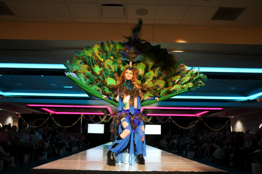 A model posing on a runway in an elaborate peacock-themed costume featuring a massive fan of peacock feathers, blue cutout pants, and black platform boots, with an audience seated in the background.