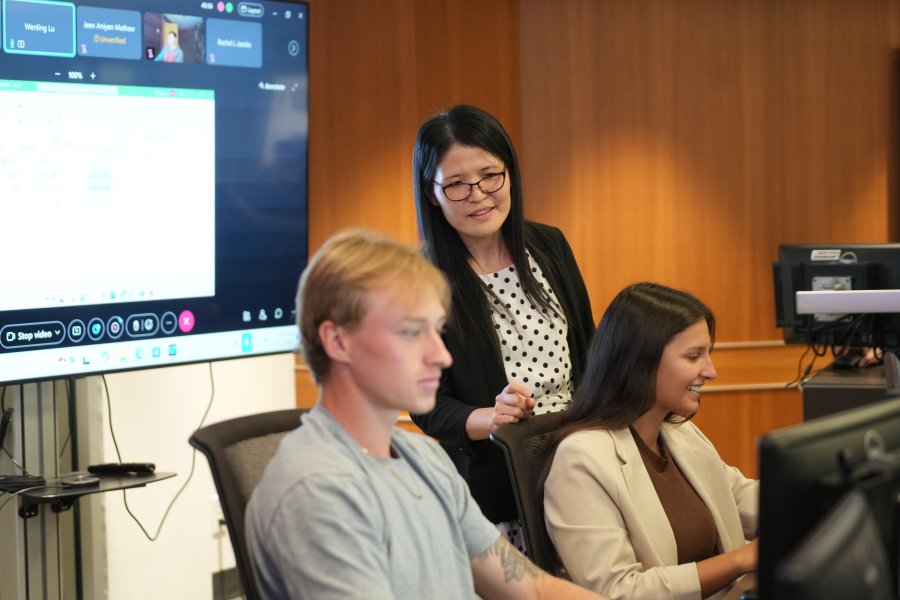 A professor speaks to a small group of students as they look at the nearby screens.