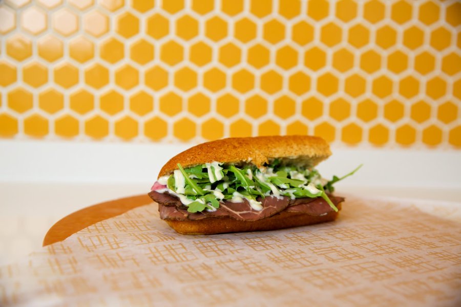 "The Buster" sandwich sits on a round wood platter with an orange tile wall in the background.