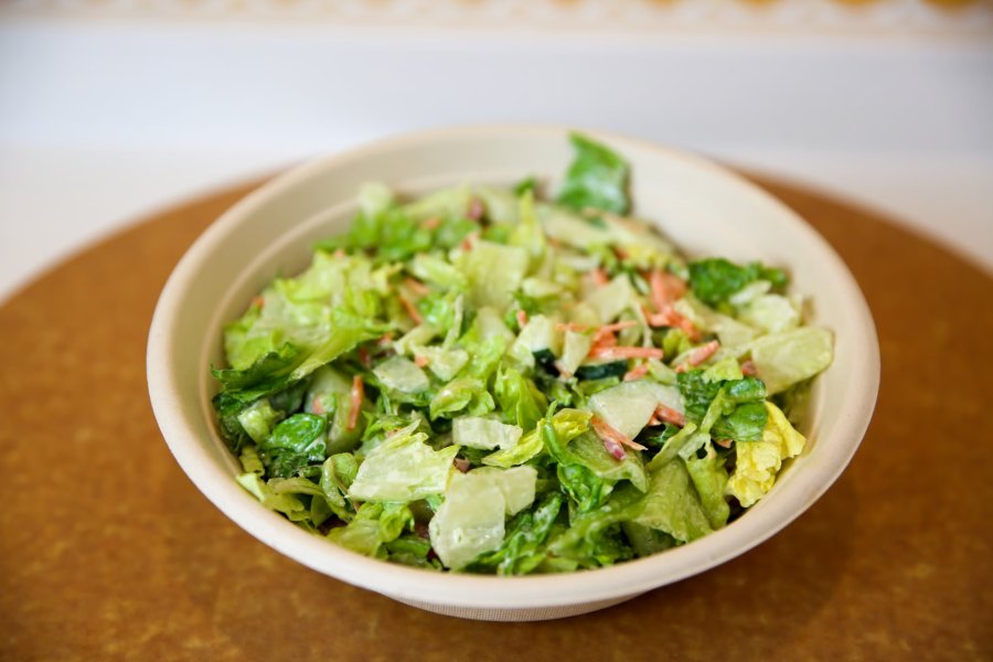 A freshly-made salad in a take-out bowl sitting on a circular wooden tray.