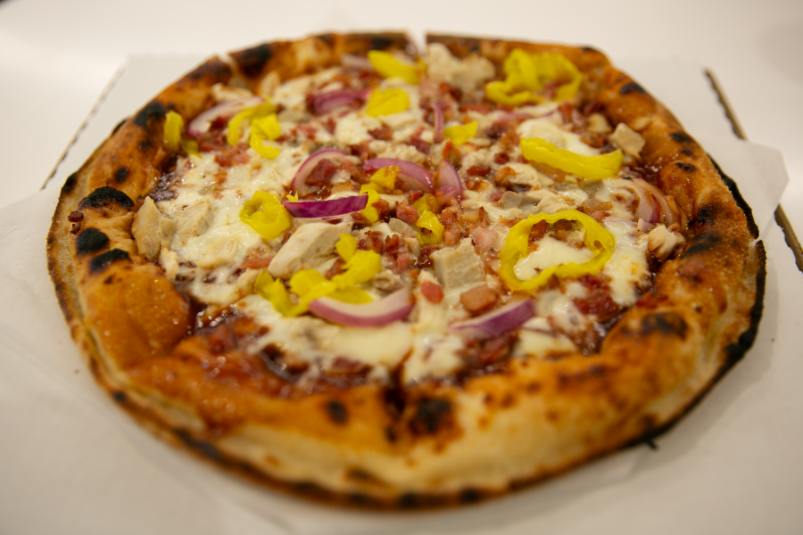 A My Pie barbecue chicken pizza with slices of red onion, crumbled bacon, and yellow banana pepper rings on a white table.