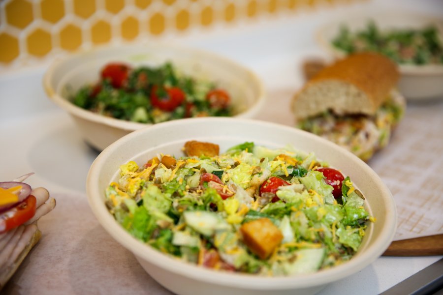 An assortment of salads, grain bowls, and sub sandwiches sitting on a white countertop.