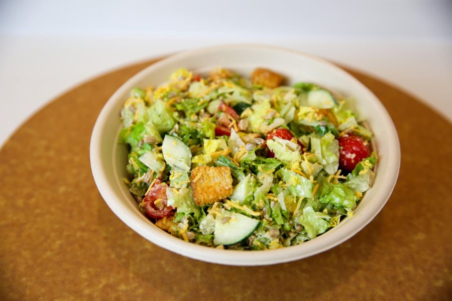 A freshly-made house salad with cucumbers, croutons, shredded cheese and tomatoes over a bed of greens in a take out bowl.
