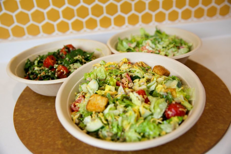 Three freshly-made salads and grain bowls in take-out bowls on a countertop.