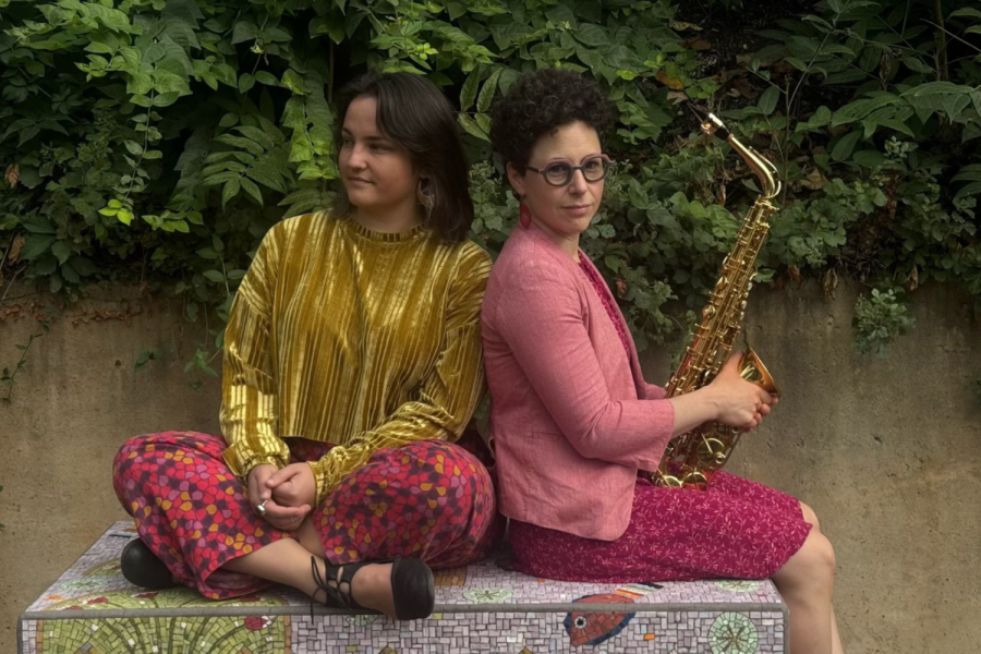 A woman in a gold top sits next to a woman in a pink top. The woman on the right is holding an alto saxophone. 