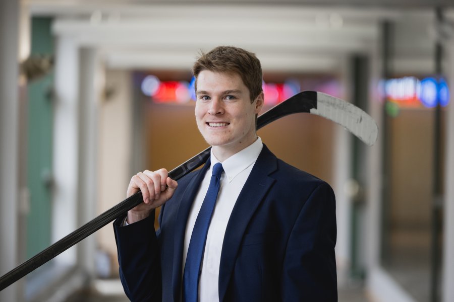 Grant Slukynsky stands in the hallway of Schneider Hall with a hockey stick over his shoulder.
