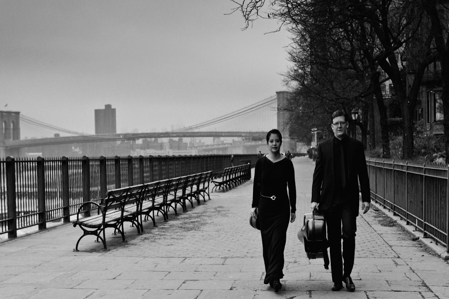 A man and a woman wearing black clothing walking along a waterfront. The woman is carrying a violin case, and the man is carrying a cello case.
