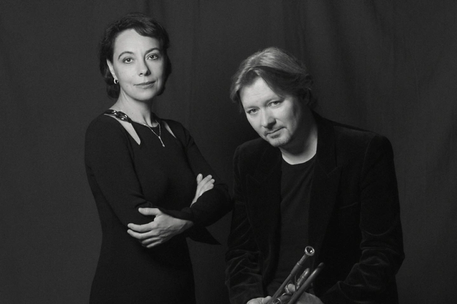 A woman and a man in dark colored clothing. The man is holding a trumpet.