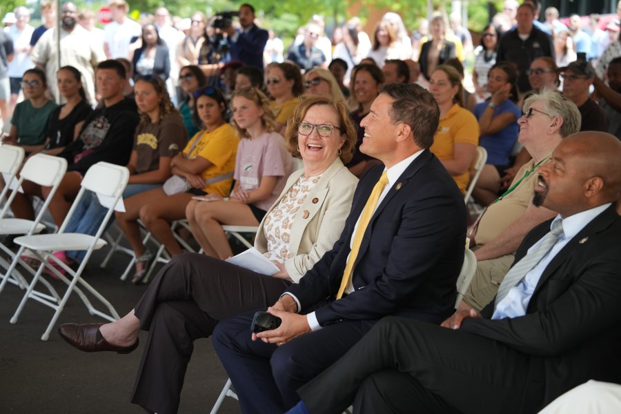 A crowd of people sits under a tent at a ribbon-cutting ceremony.