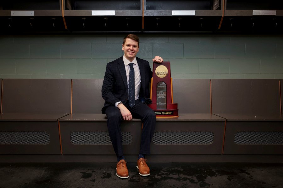 Travis Culhane, wearing a suit, sits next to a sports trophy.