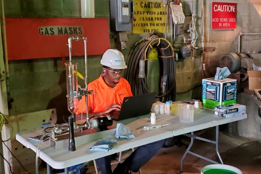 JustOne Crosby, wearing an orange shirt and white hard hat and safety glasses, sits at a laptop in an industrial setting.