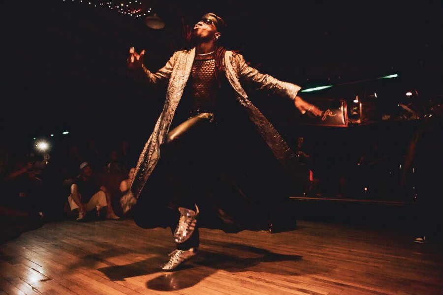 A man dances on a dance floor in a long, silver coat and silver shoes.