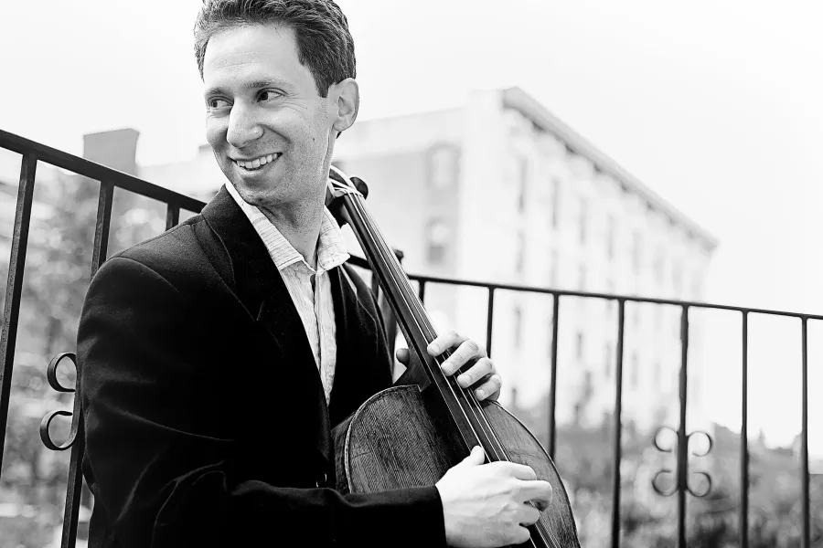 A smiling man in a dark suit playing a cello.