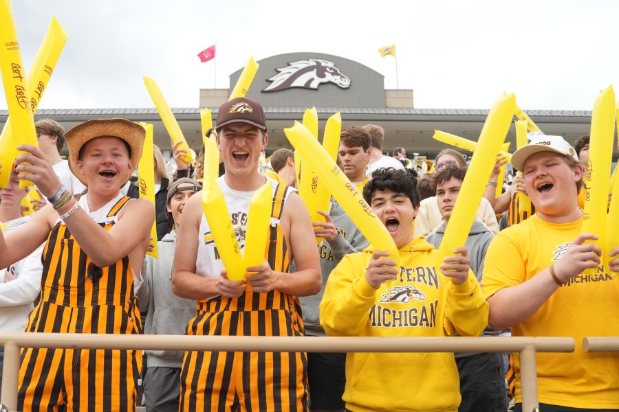 A crowd of enthusiastic Western Michigan Broncos fans cheering in the stands, holding yellow inflatable thundersticks. Some fans are wearing yellow and black striped overalls and school apparel, with the Broncos horse logo visible on a stadium structure in the background.