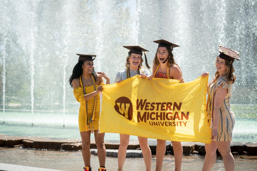 Four graduates wearing graduation caps are standing in front of a large water fountain, smiling and laughing as they hold up a yellow Western Michigan University flag.