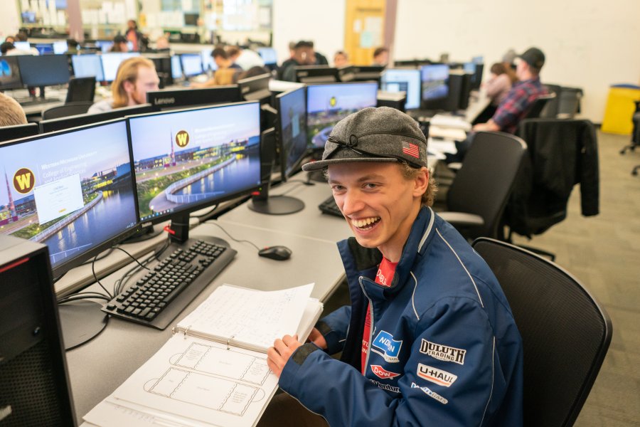 A College of Engineering student sits at a bank of monitors, smiling. He is wearing a hat and jacket. 