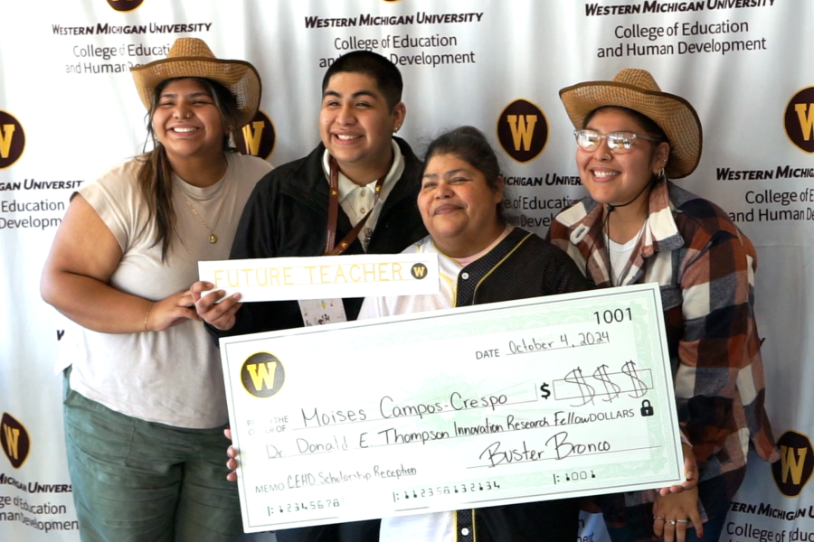 Four smiling individuals posing together in front of a Western Michigan University backdrop, with two wearing cowboy hats and holding a novelty oversized scholarship check presented to Moises Campos-Crespo, alongside a "Future Teacher" sign. 