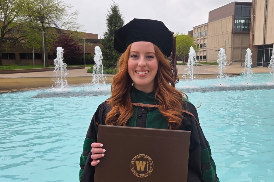 Honors alumna Serena Karlov stands in full regalia, holding her diploma folder, in front of Miller Fountain.