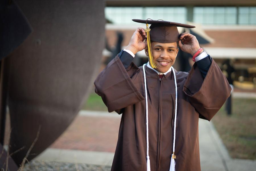 A student, dressed in a grad cap and gown, adjusts their tassel. 