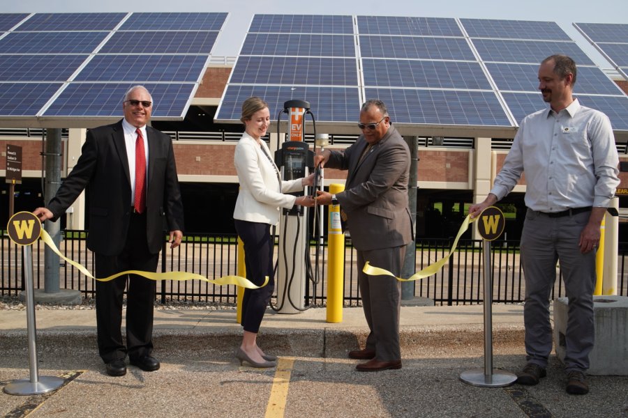 University officials use a pair of large scissors to cut a yellow ribbon in front of an electric vehicle charging station.