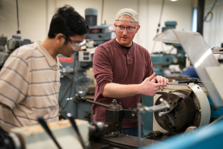 Two men in a machine shop stand at a large metal lathe, with one man pointing and explaining its operation while the other watches.