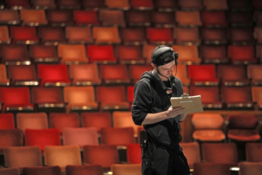 Young man wearing black and a headset stands in a theatre with clipboard.