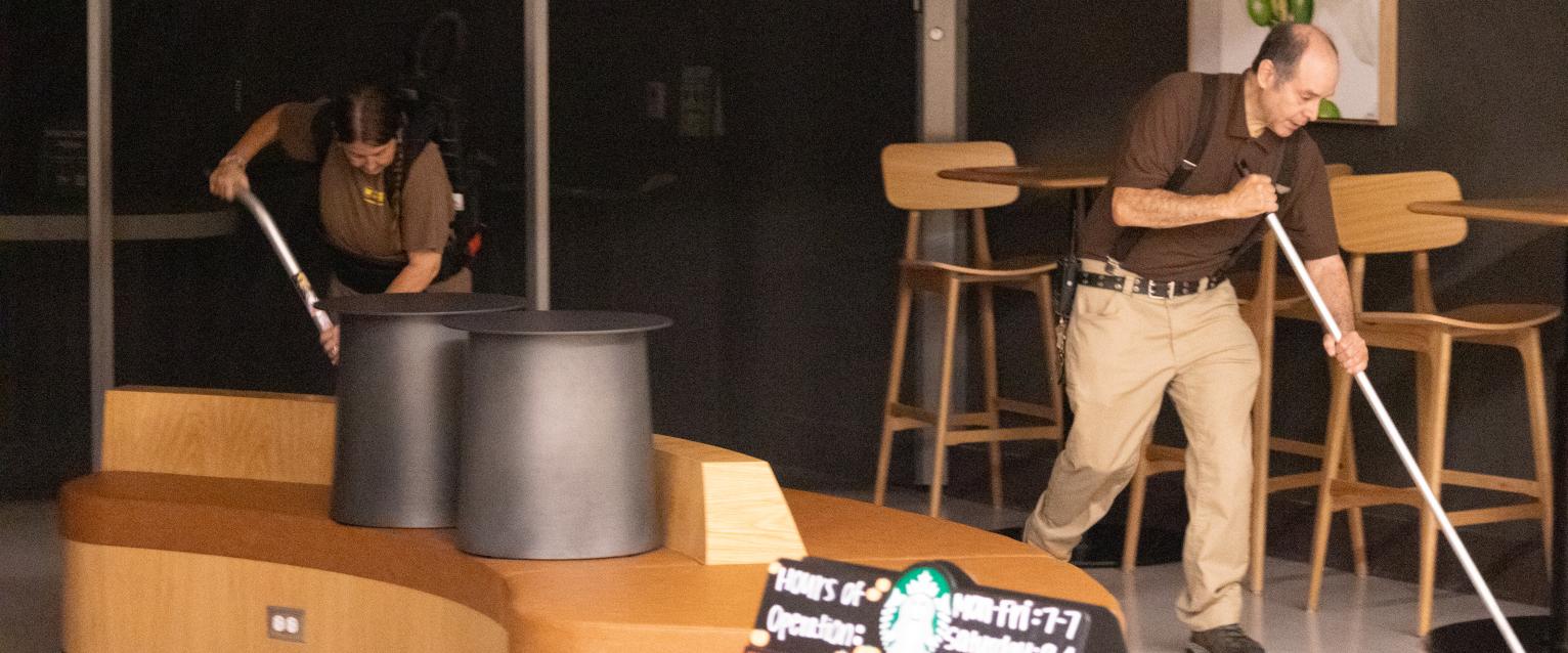 Two custodians are seen cleaning the entry seating area of Starbucks at the Student Center. One is vacuuming and one is mopping.