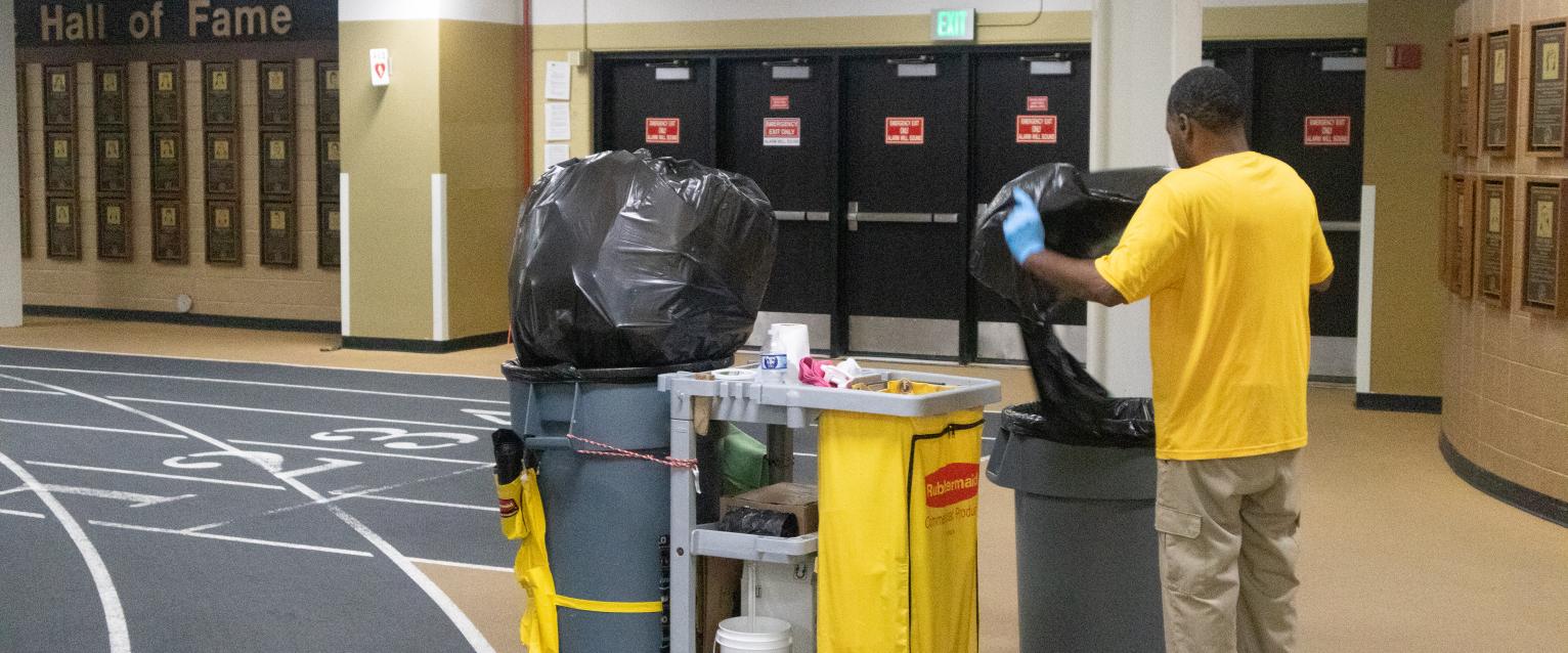 A custodian is seen changing the trash bag for a trash bin near the entrance of University Arena.