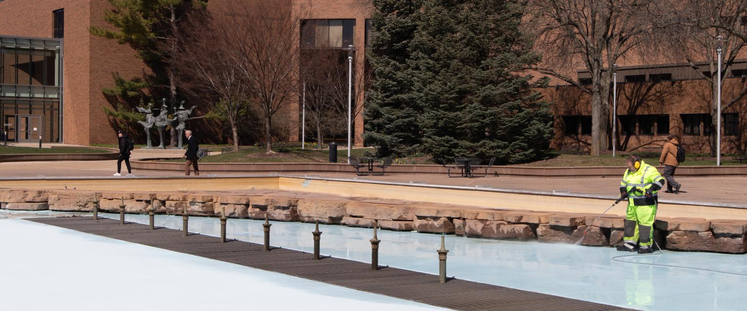 A custodian is seen in a neon green work outfit, power washing Miller Fountain.