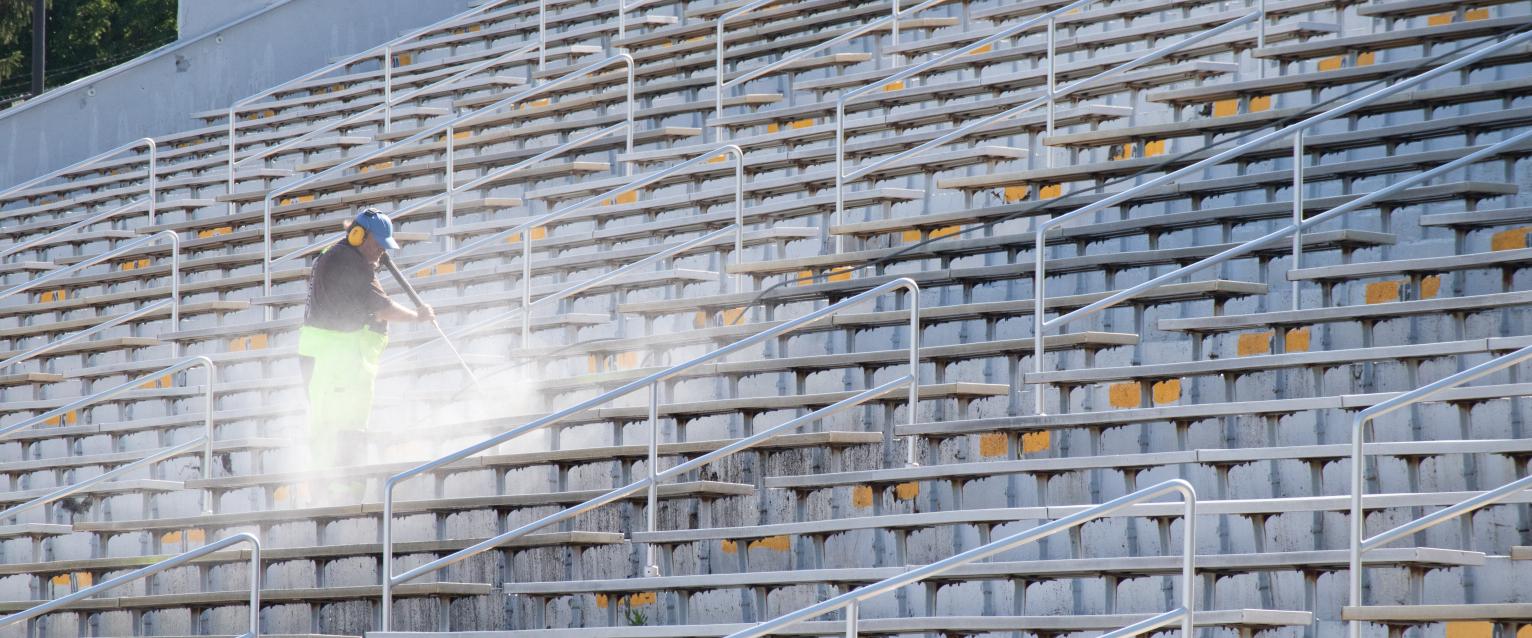 Custodial employee seen power washing bleachers while wearing a brown shirt, neon yellow pants and ear protection.