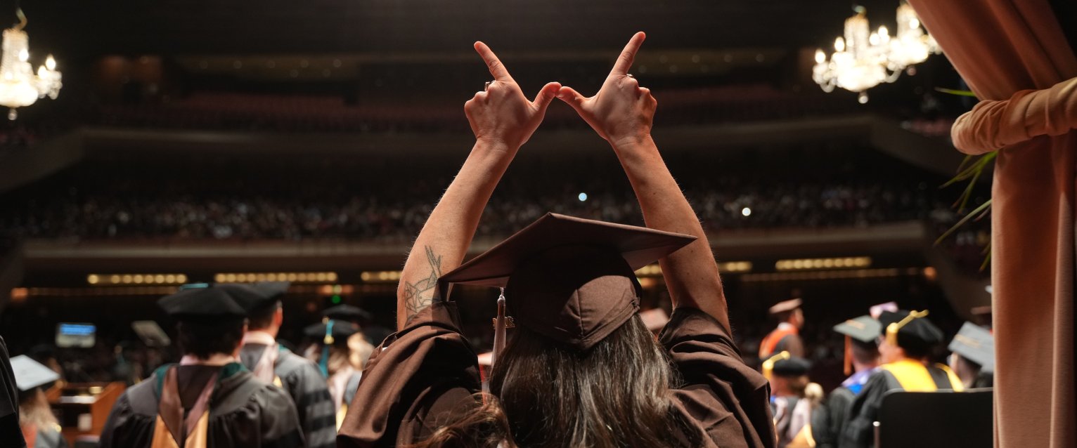 A student creating a W with her hands on the stage of Miller Auditorium at WMU commencement.