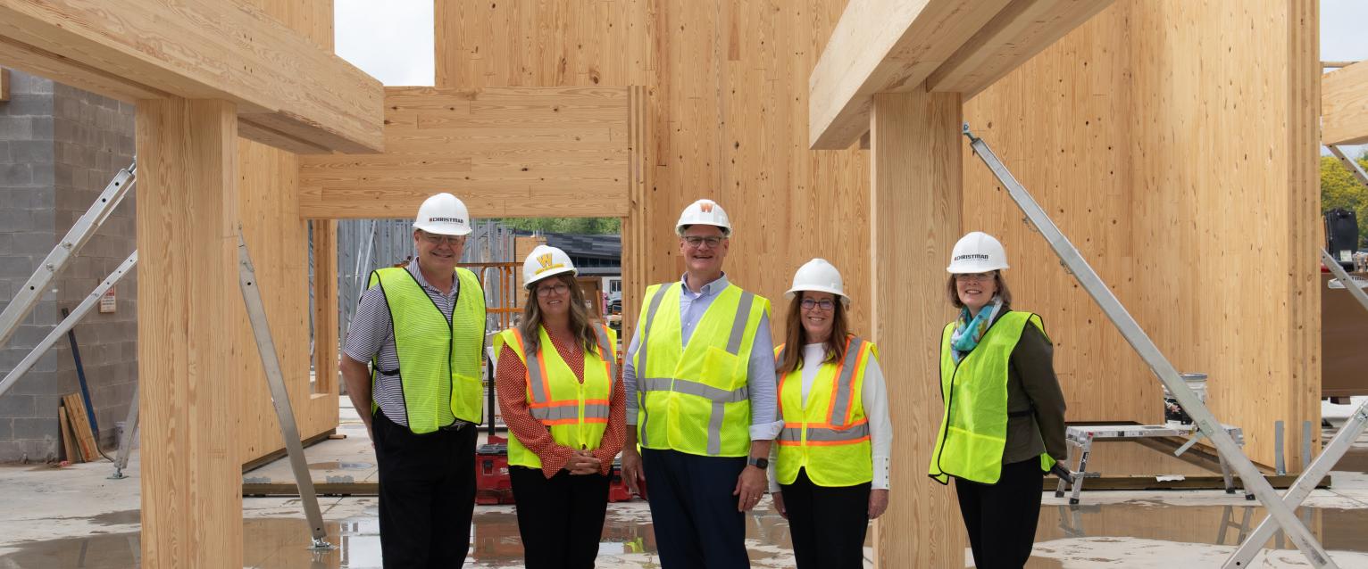 A group of five employees from WMU stand in front of one of the mass timber walls on the construction site of the new residence hall. All five are wearing safety vests, white hard hat and safety glasses.