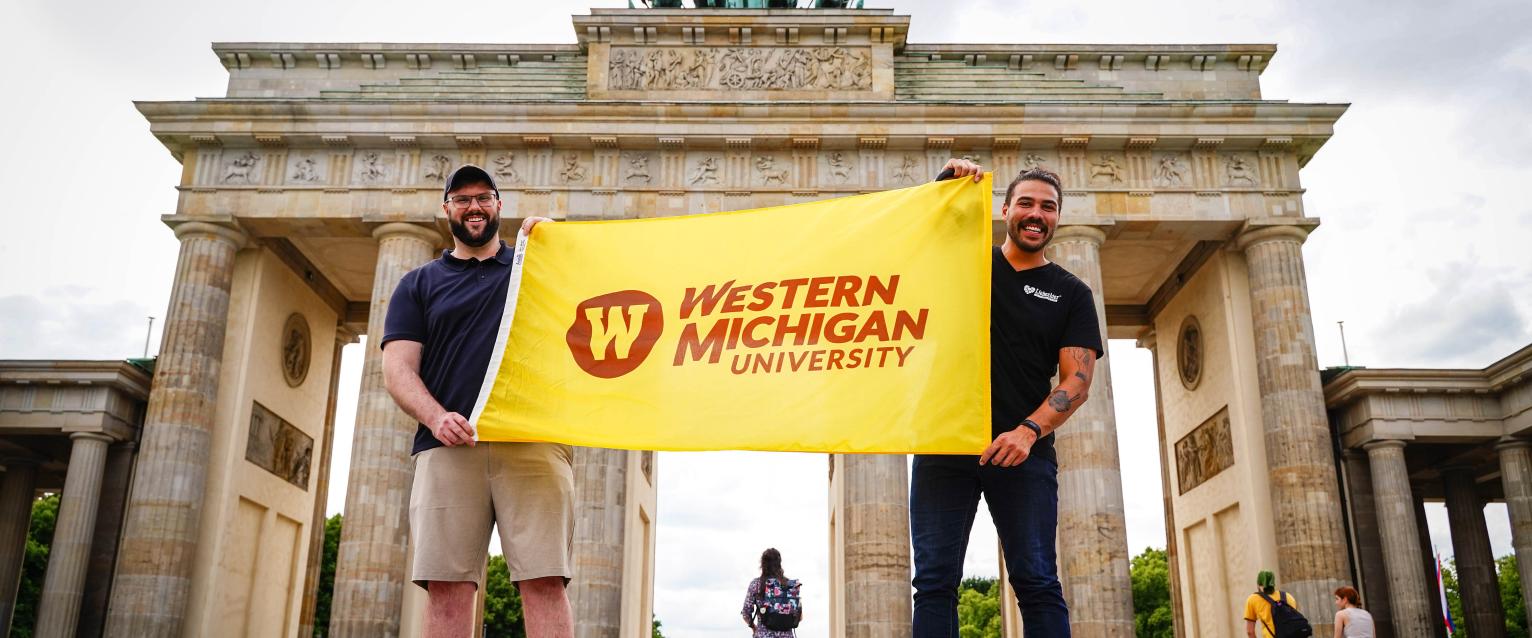 Two people holding the WMU flag in front of Brandenburg Gate in Berlin.