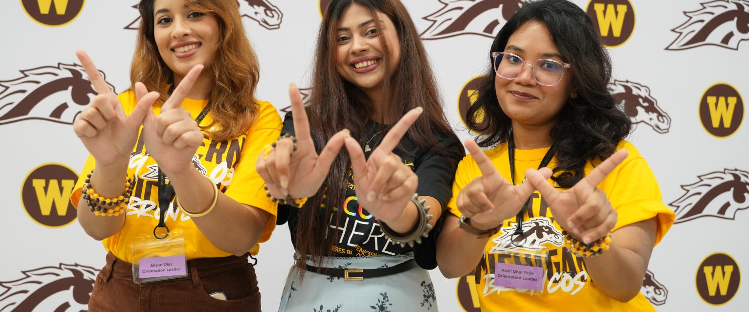 Three students smiling at the camera and making the W sign in front of a WMU logo backdrop.