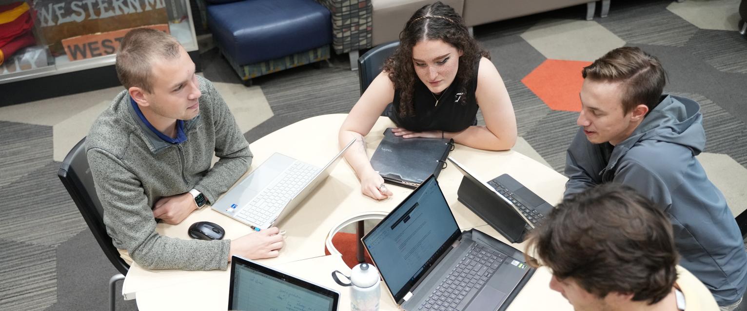 Students with laptops and backpacks working at table.