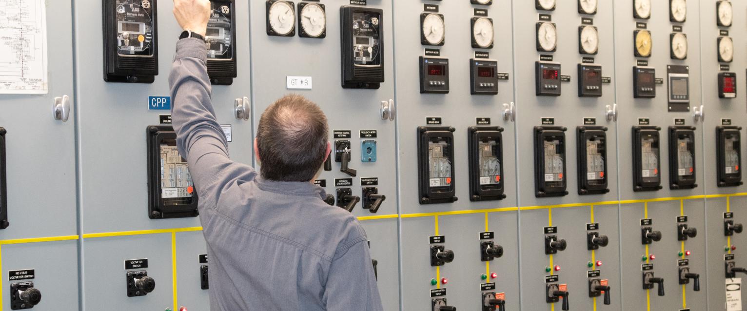 Power plant employee is facing a wall of switches and gauges, pointing at a gauge at the top of the panel.