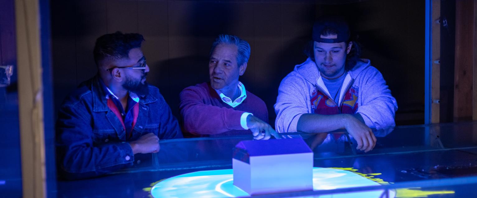 Three men standing over a small house, looking at the wind around it. 