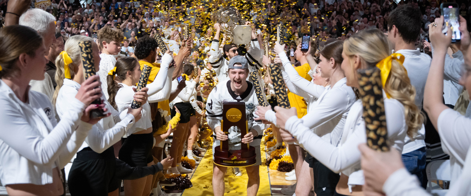 A crowd cheers while a hockey player holds a national championship trophy.