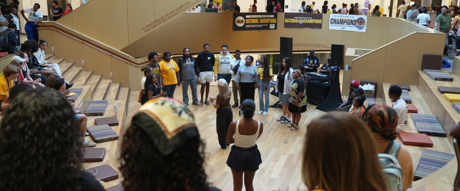Multiple students surround the Gathering Stairs in the Student Center to listen to listen to a chorus of diverse singers singing a song.