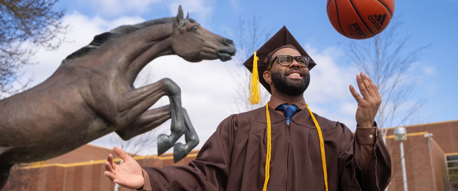 Ethan Cheatham in his cap and gown balancing a basketball on one finger in front of the Bronco statue and Reed Fieldhouse.