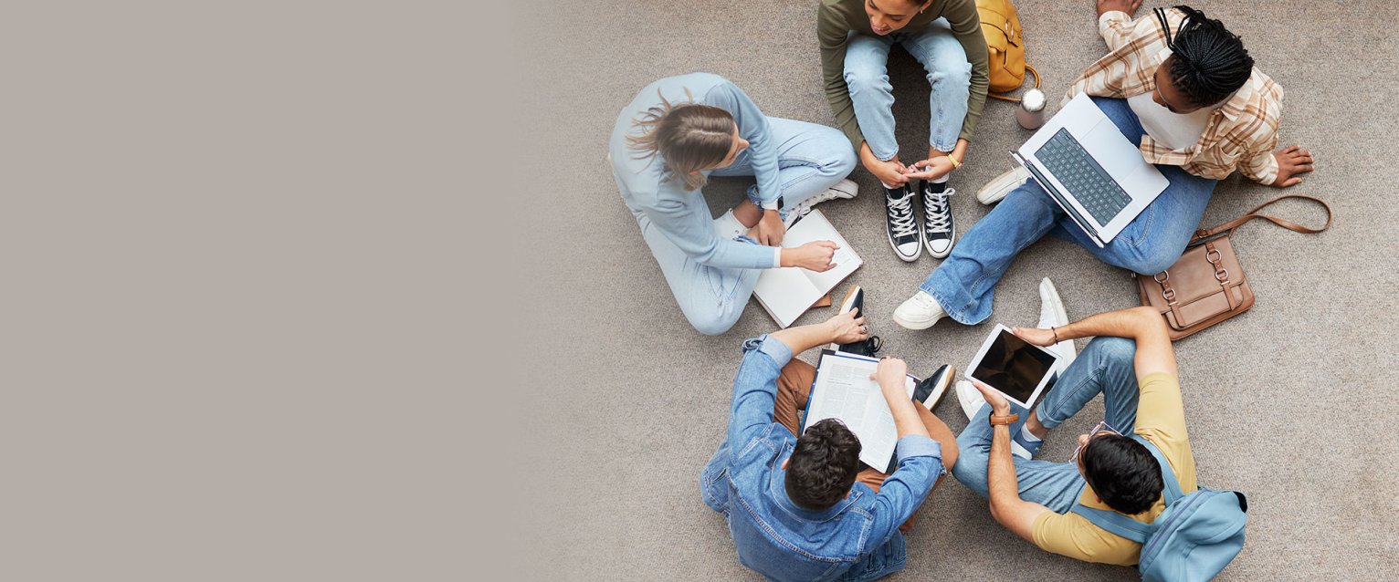 Group of students sitting on a carpeted floor in a circle with various devices such as computers and tablets.