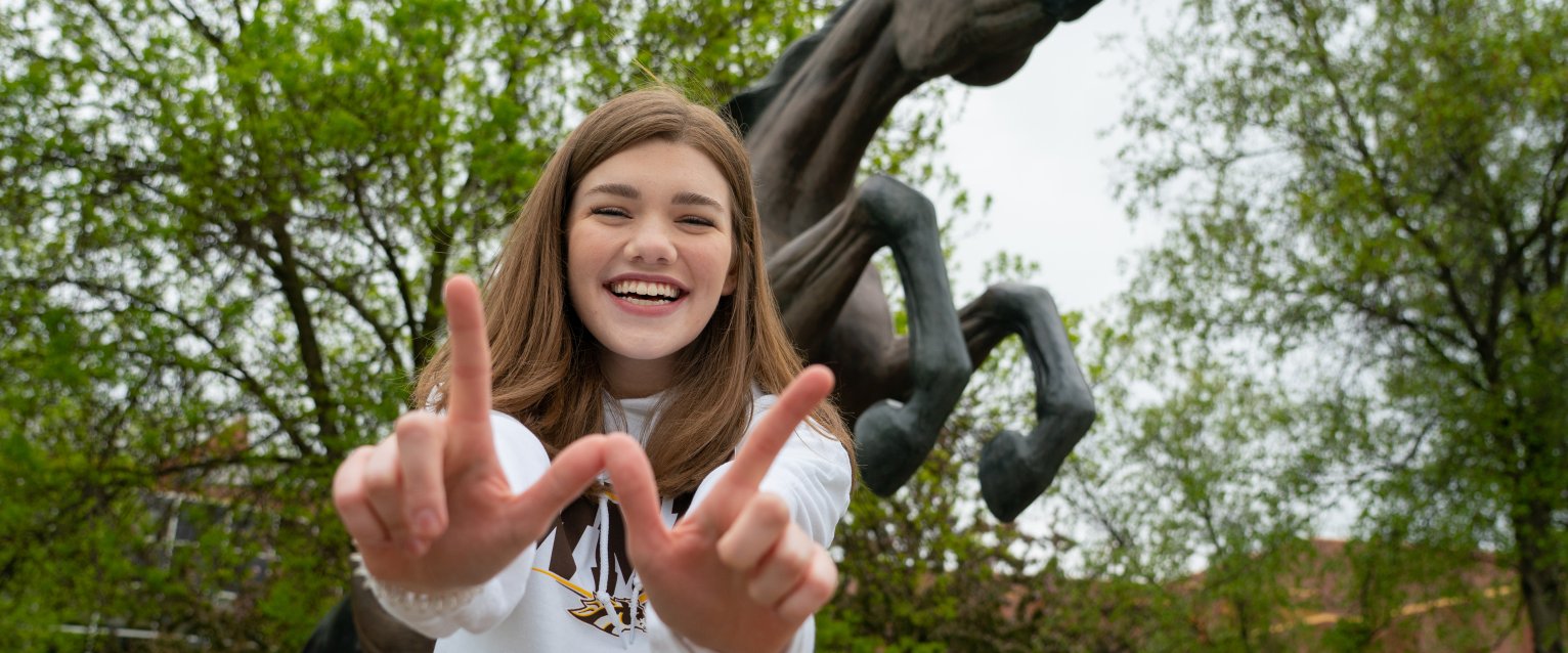 Student smiling at the camera, making a "W" with their hands, standing in front of the Bronco statue.