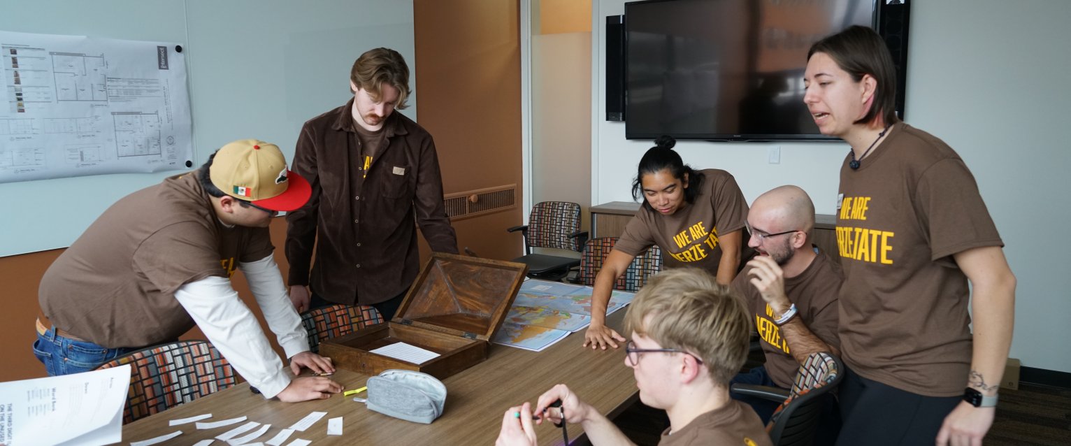 Students around a table solving an escape room puzzle in Ellsworth Hall, at Merze Tate Day Celebrations 2025.