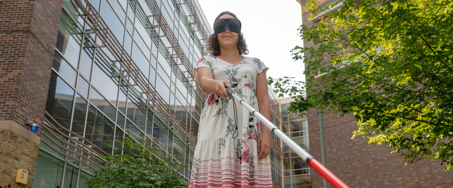 Female blindnessa nd low vision studies student navigating outdoors with a white can and eye mask