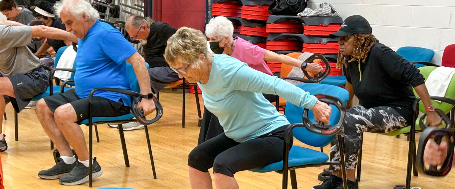 Participants of the Active Aging fitness class sitting in a row of chairs swinging their arm back with hand weights.
