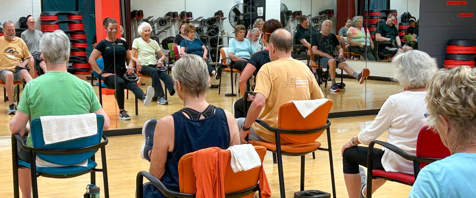 A room full of active aging participants facing a workout mirror sitting in chairs kicking their legs forward.