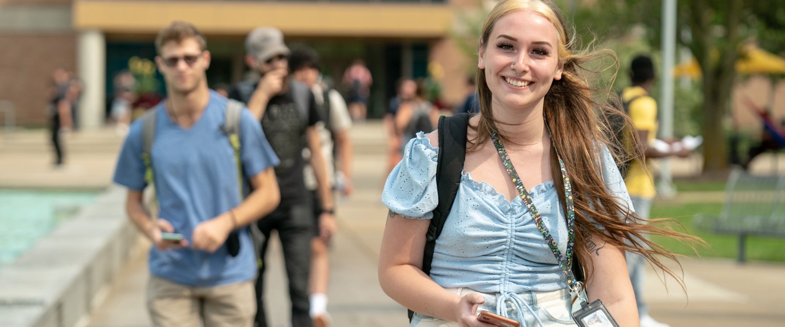 A student happily walking through WMU Main Campus with her ID lanyard and phone.