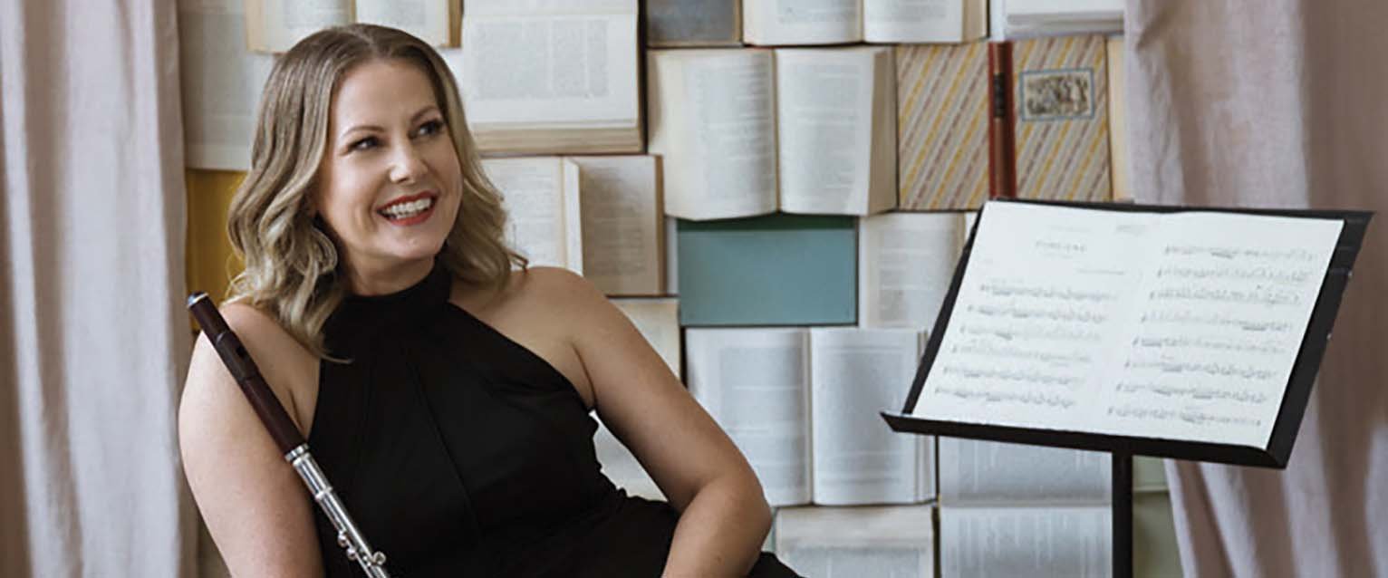 photo of person holding a flute and sitting in front of a wall with open books on display